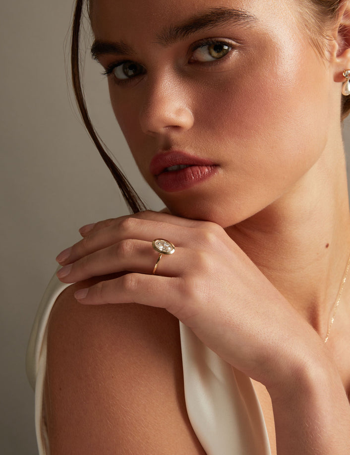 Model. A close-up lifestyle shot of a woman wearing a delicate gold ring with an oval diamond on her finger, posed against a soft neutral background.