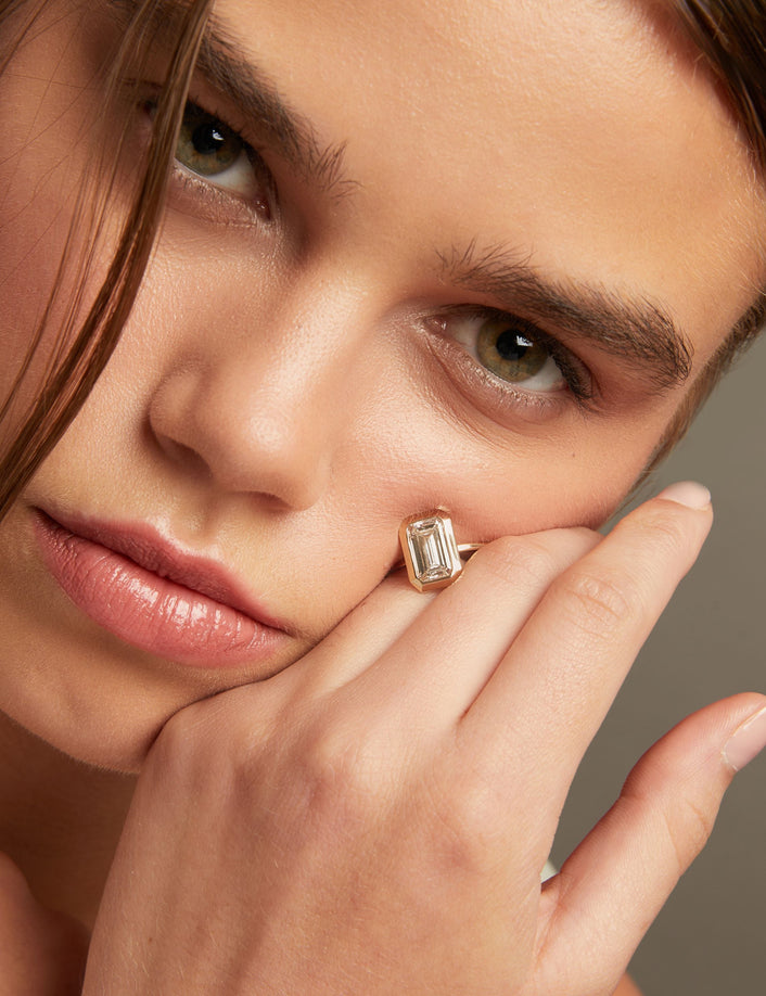 Model. Close-up of a woman with an oval diamond bezel ring resting on her cheek and hand, against a neutral gray background.