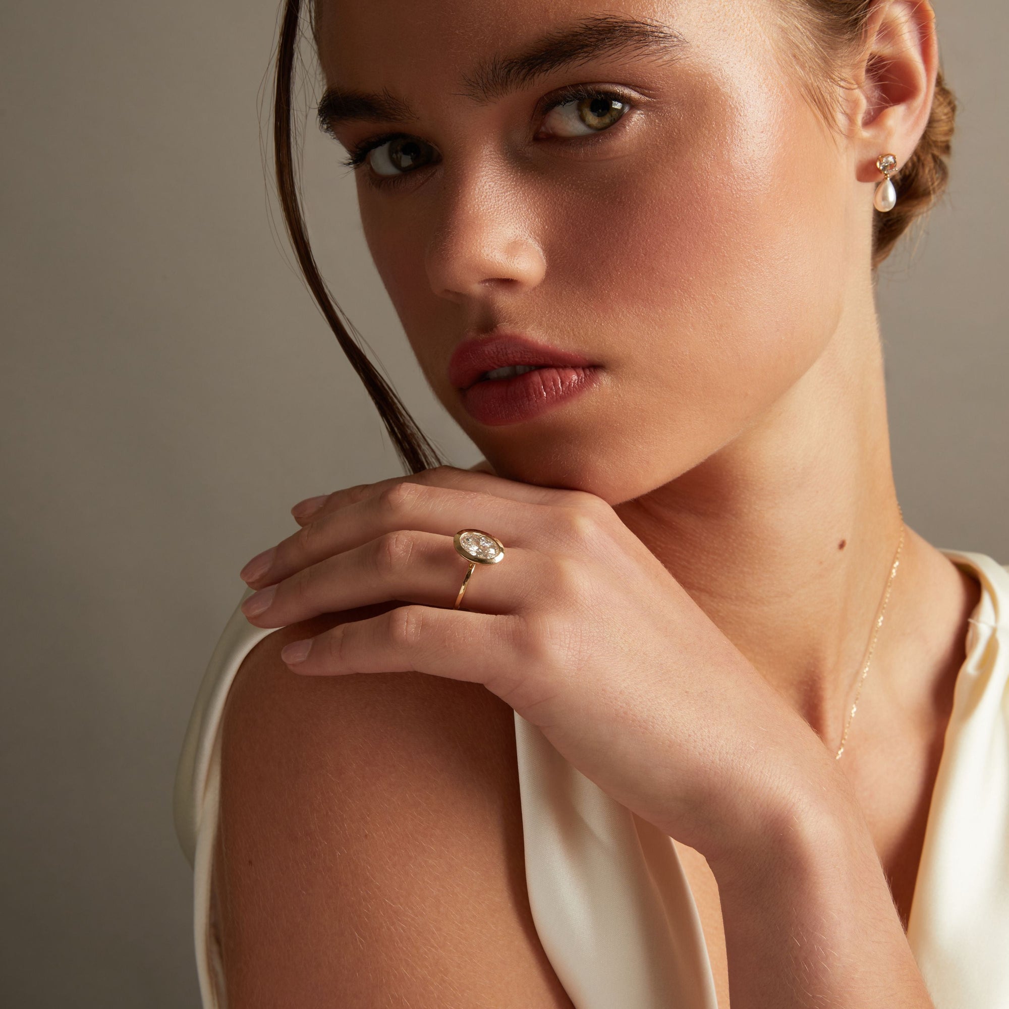 Model. A close-up lifestyle shot of a woman wearing a delicate gold ring with an oval diamond on her finger, posed against a soft neutral background.