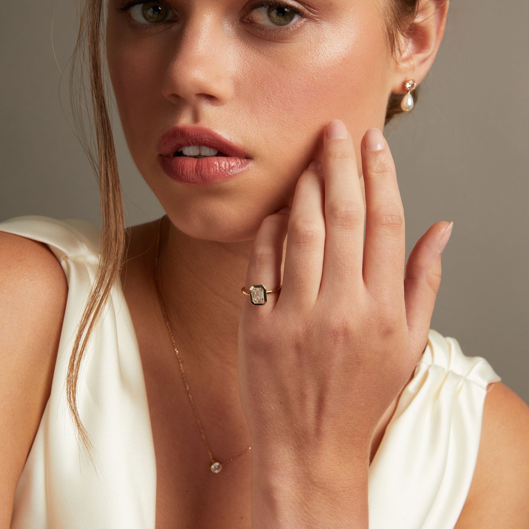 A close-up lifestyle shot of a woman wearing a gold ring with an emerald diamond on her finger, along with matching pearl drop earrings and a delicate gold necklace.