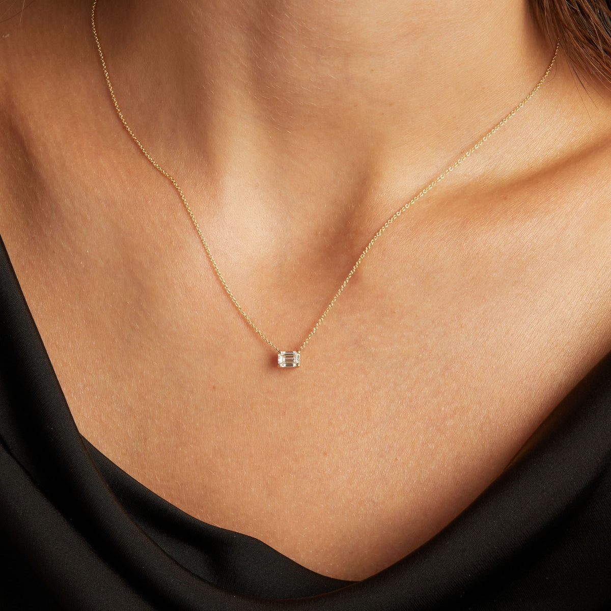 Model. Close up of woman wearing a black top with an emerald diamond solitaire necklaces on a beige background.
