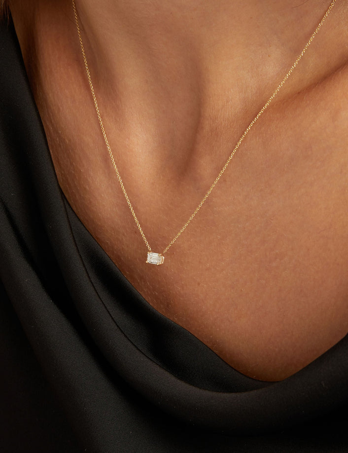 Model. Close up of woman wearing a black top with an emerald diamond solitaire necklaces on a beige background.