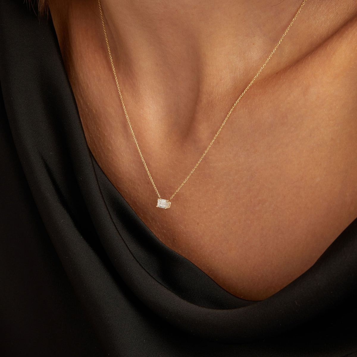 Model. Close up of woman wearing a black top with an emerald diamond solitaire necklaces on a beige background.