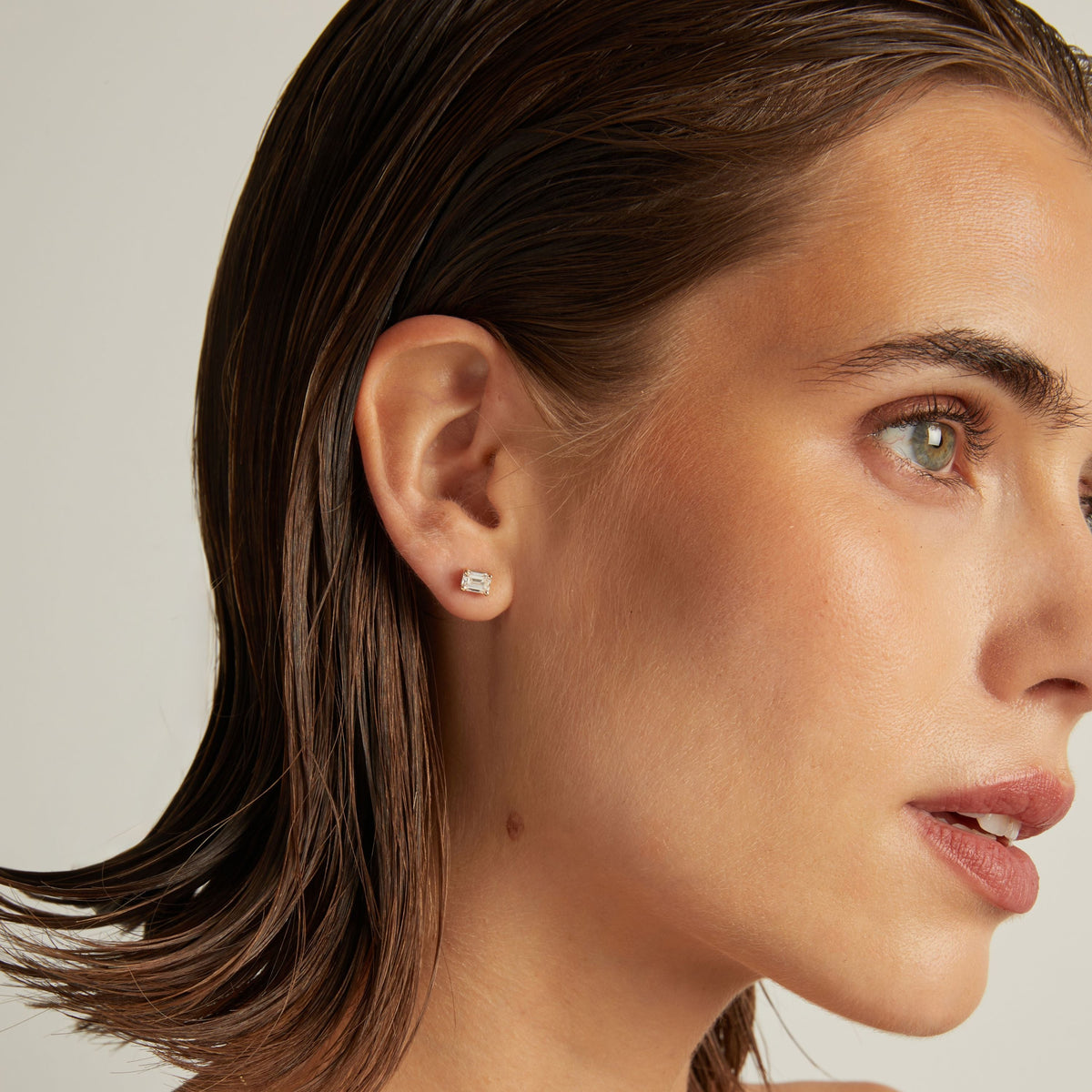 Close up of woman wearing an emerald shaped diamond stud earring on a beige background.