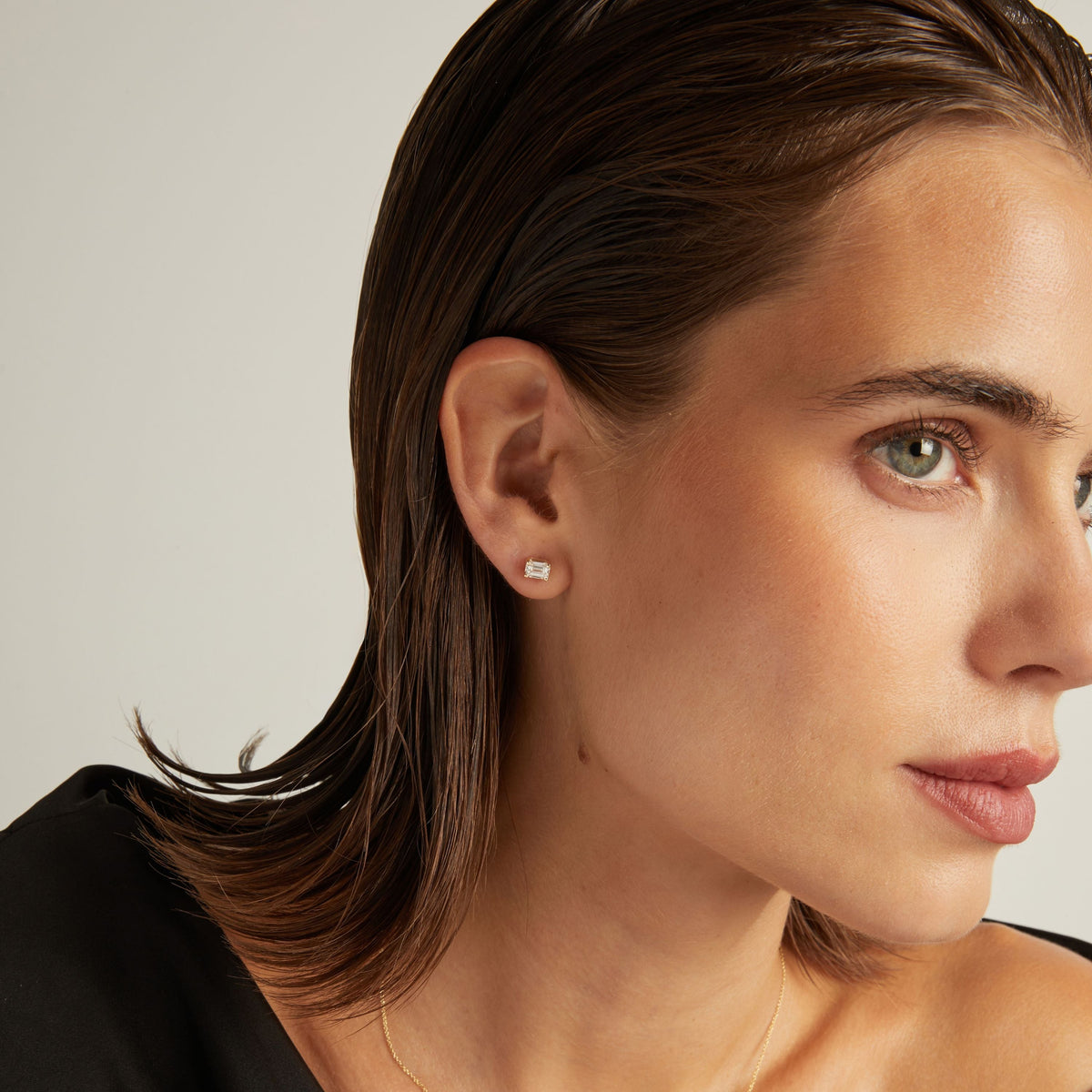 Model. Close up of woman wearing an emerald shaped diamond stud earring on a beige background.