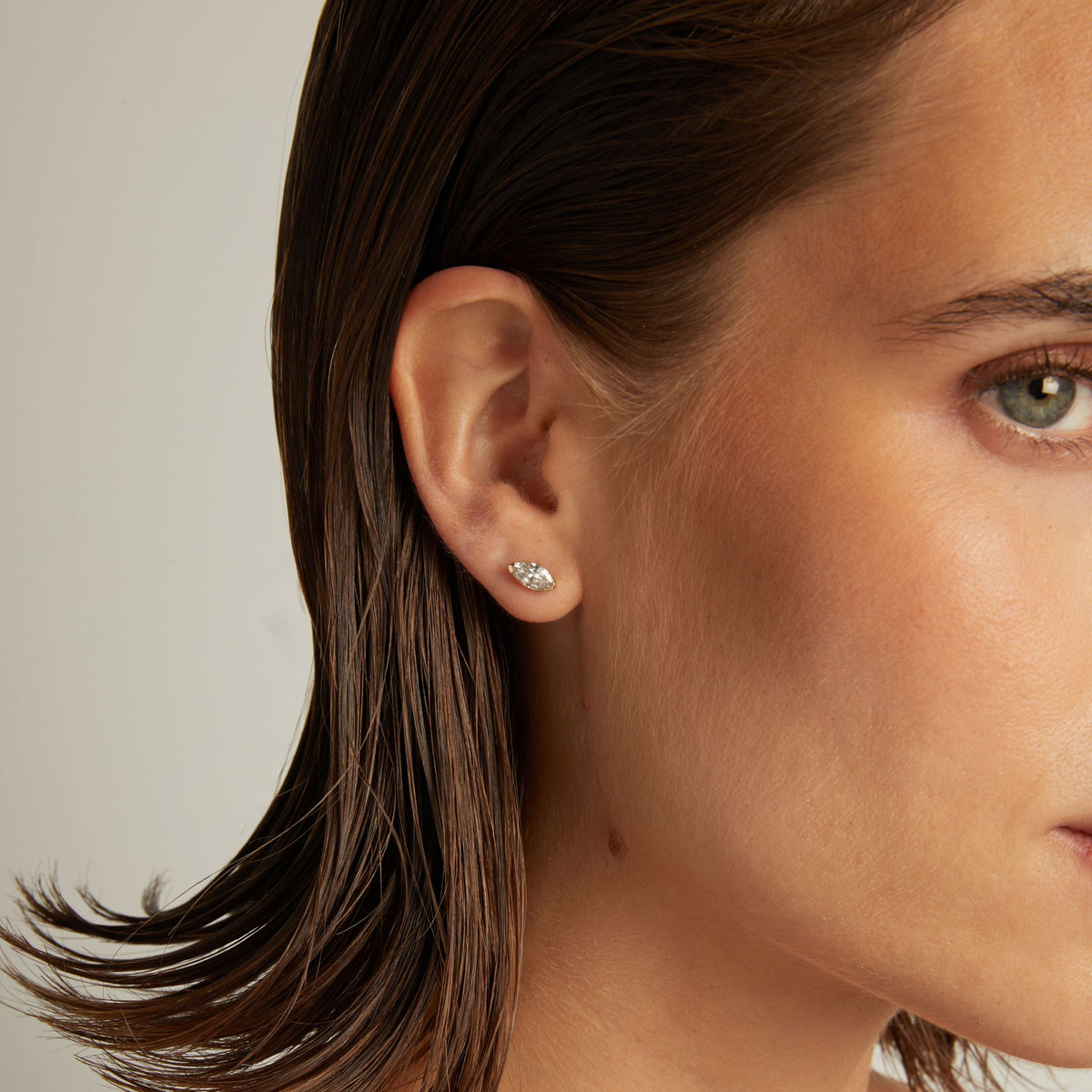 Model. Close up of woman wearing a marquise shaped diamond stud earring on a beige background.