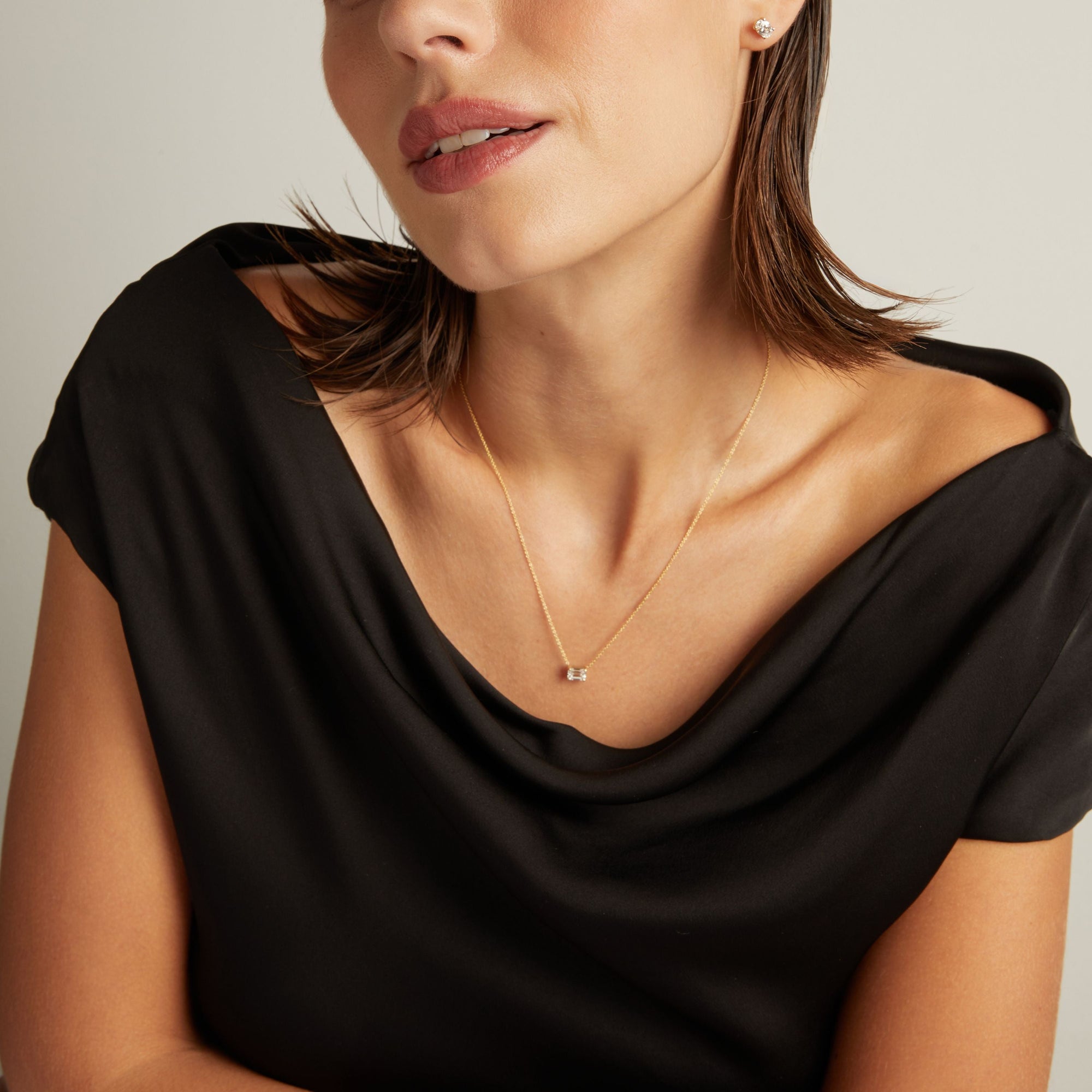 Model. Close up of woman wearing a black top with an emerald shaped diamond solitaire necklaces on a beige background.