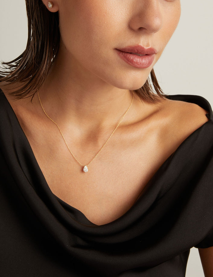 Model. Close up of woman wearing a black top with a pear shaped diamond solitaire necklaces on a beige background.