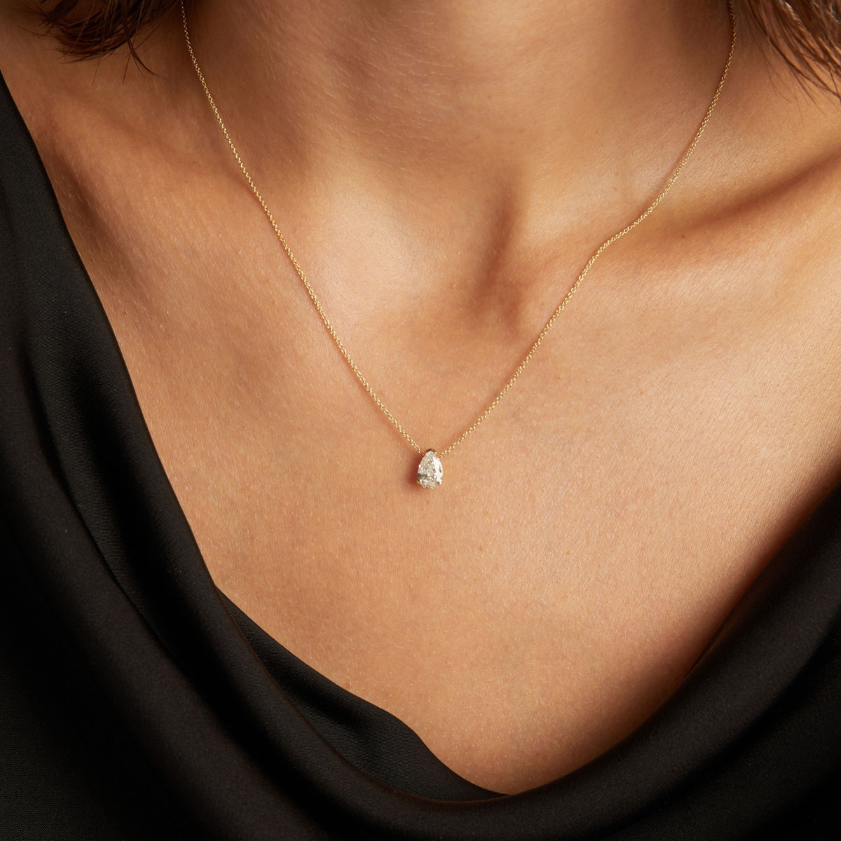 Model. Close up of woman wearing a black top with a pear shaped diamond solitaire necklaces on a beige background.