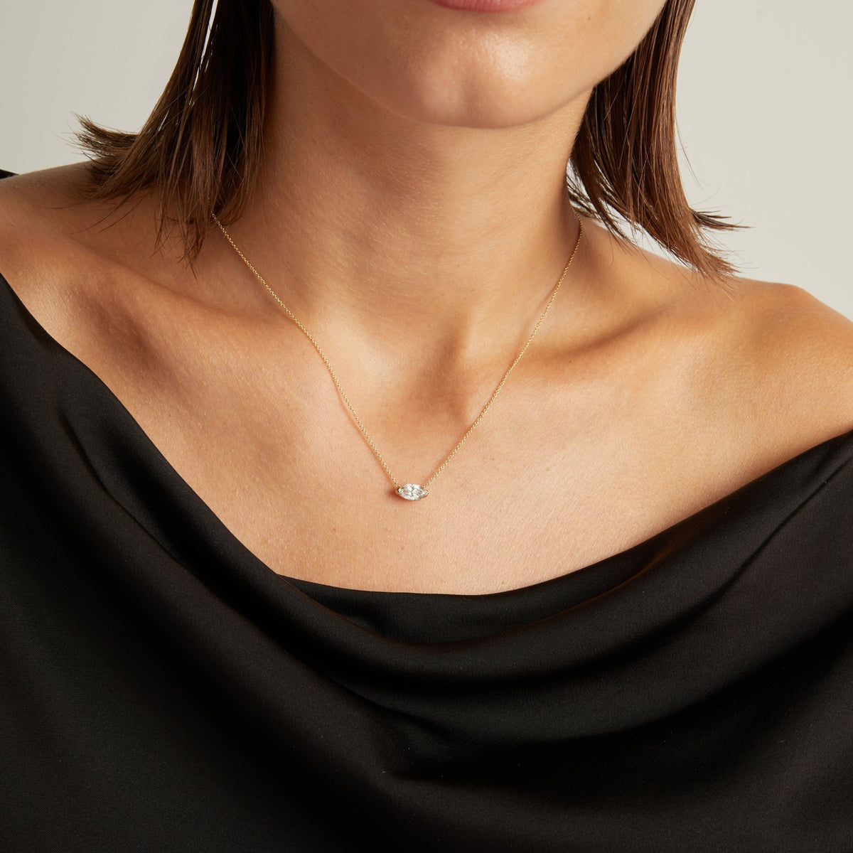 Model. Close up of woman wearing a black top with a marquise shaped diamond solitaire necklaces on a beige background.