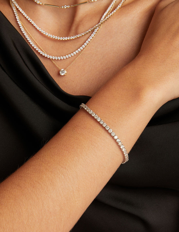 Model. Close up of a woman wearing a black top with stacked diamond necklaces and a diamond tennis bracelet on beige background.