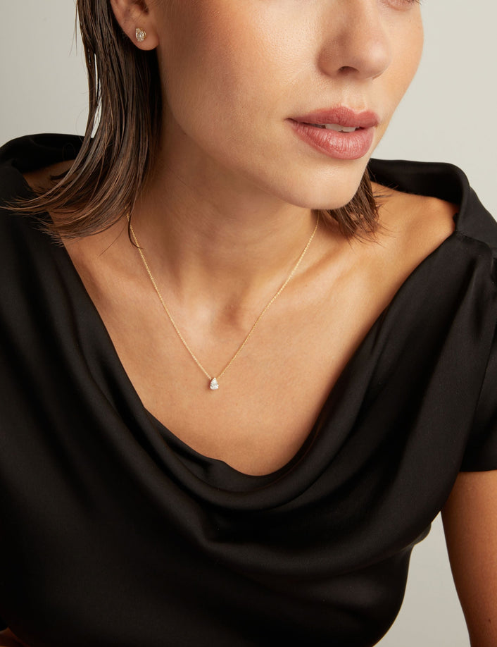 Model. Close up of woman wearing a black top with a pear diamond solitaire necklaces on a beige background.
