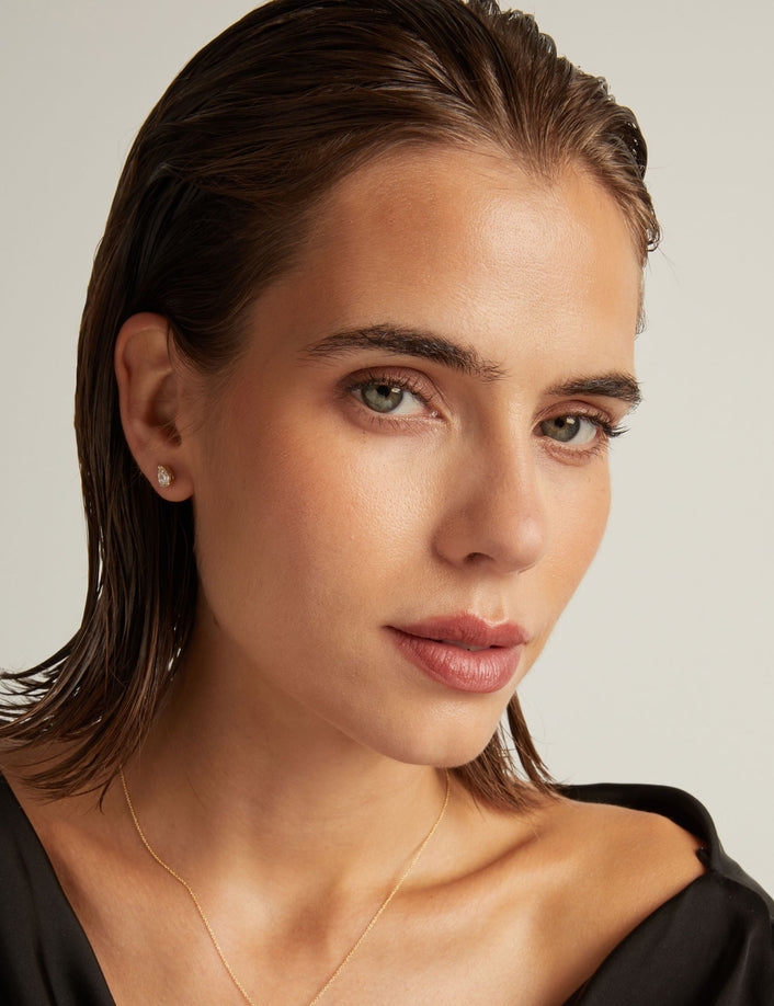 Model. Close up of woman wearing a pear diamond stud earring on a beige background.