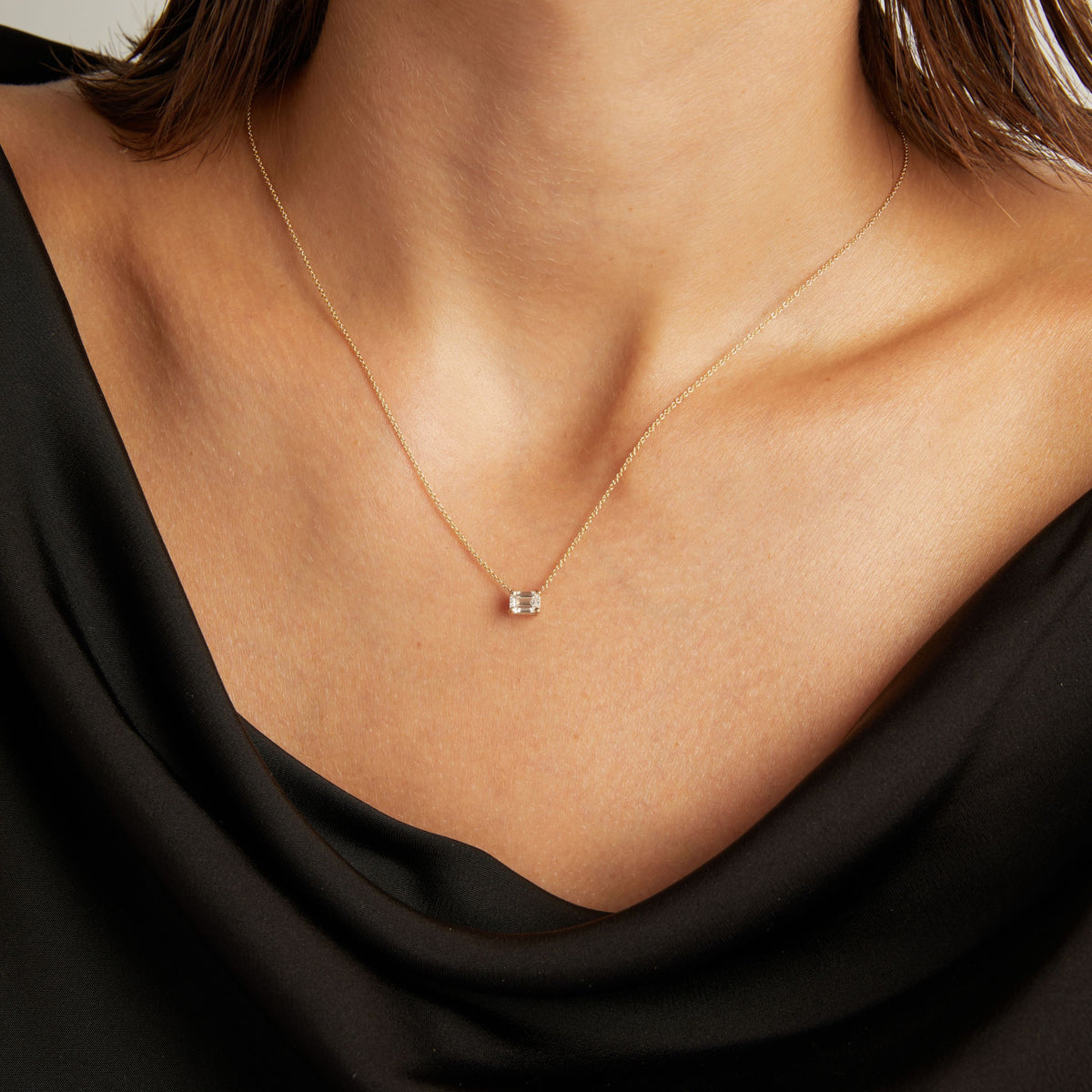 Close up of woman wearing a black top with an emerald diamond solitaire necklaces on a beige background.