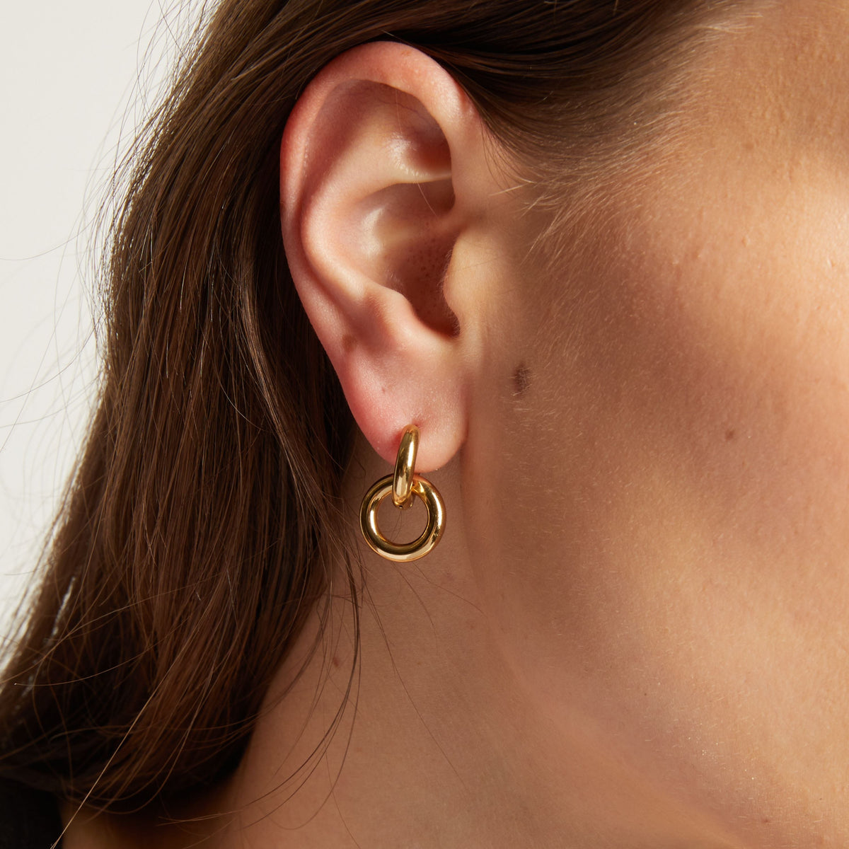 Model. Close up of a person wearing a gold hoop earring with a plain background.