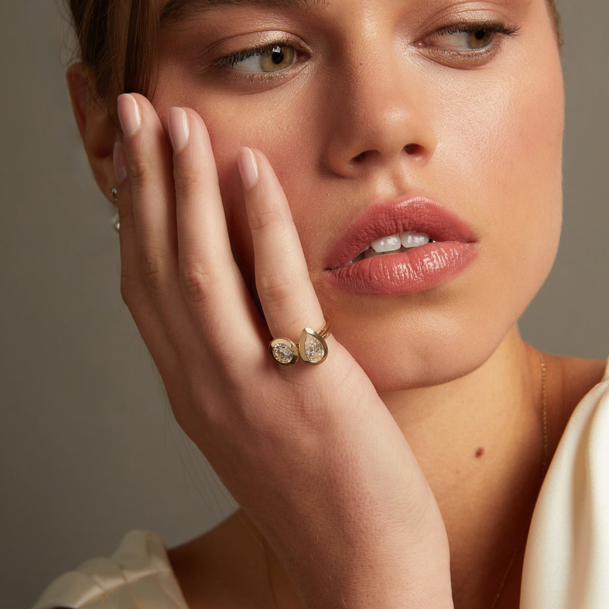 Model. A close-up lifestyle shot of a model wearing two gold rings with a pear and round diamond, with matching pearl drop earrings visible on her ear lobe.