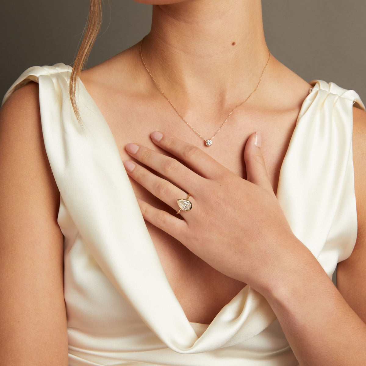 A close-up lifestyle shot of a woman's hand wearing a gold ring with a pear bezel-set diamond. She is wearing a cream satin top against a neutral gray background.