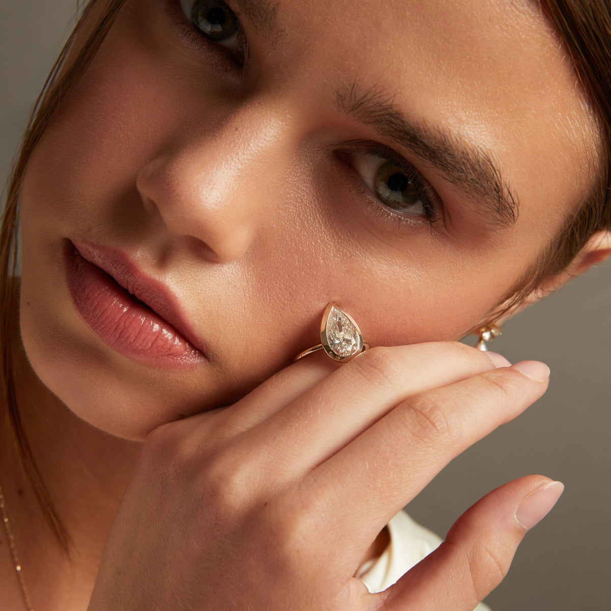 Model. A close-up lifestyle shot of a model resting a gold ring with a pear-shaped diamond on her hand and face.