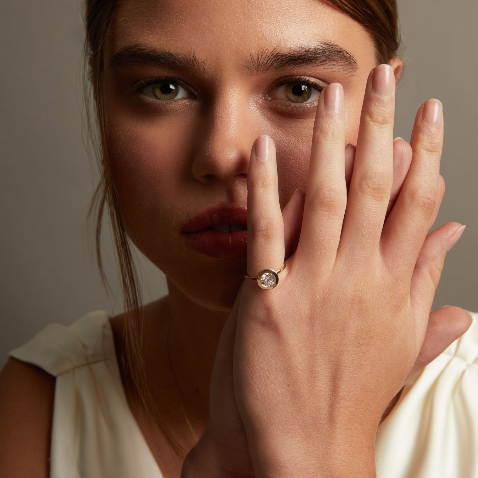 Model. A close-up lifestyle shot of a woman wearing a delicate gold ring with a small round clear gemstone on her finger, posed against a soft neutral background.