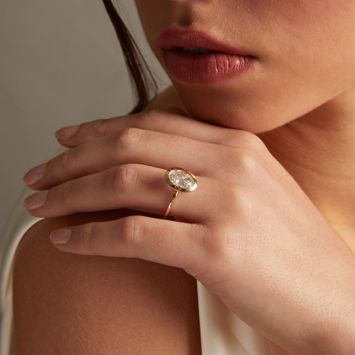 Model. A close-up lifestyle shot of a woman wearing a delicate gold ring with an oval diamond on her finger, posed against a soft neutral background.