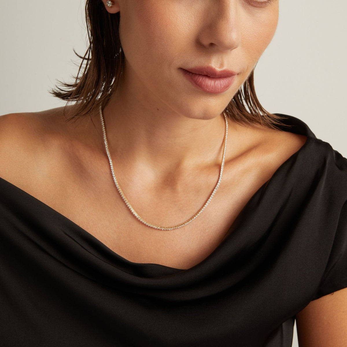 Close up of a woman in a black top wearing a diamond tennis necklace on beige background.