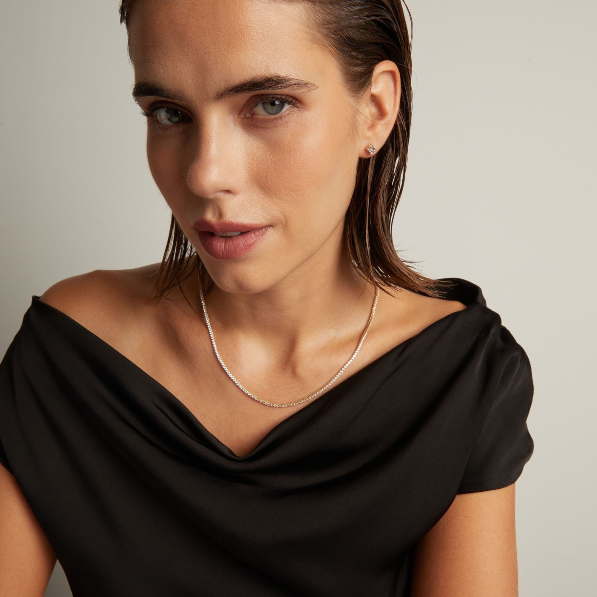 Close up of a woman in a black top wearing a diamond tennis necklace on beige background.