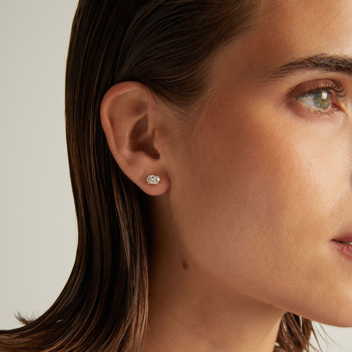 Model. Close up of woman wearing an oval diamond stud earring on a beige background.