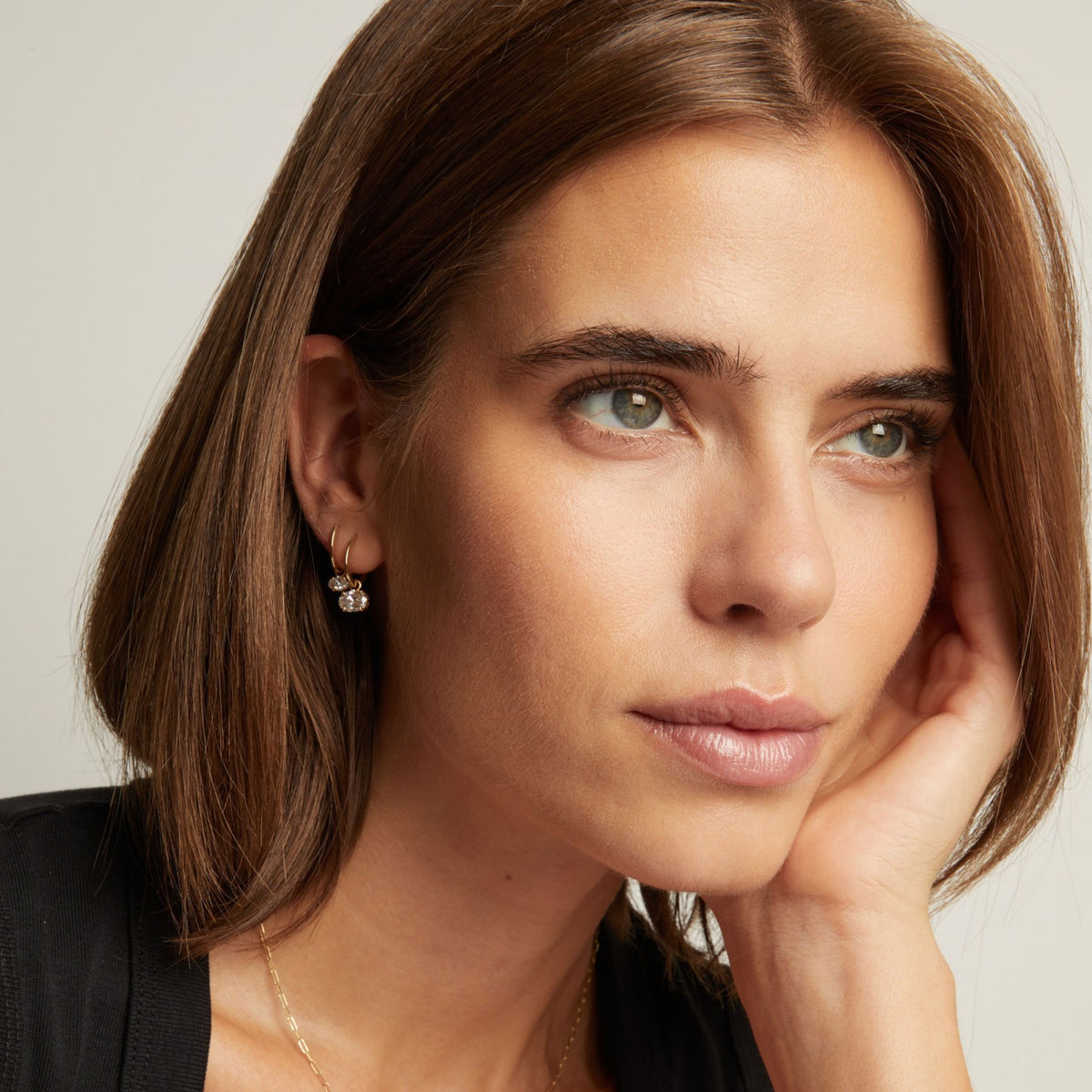 Model. Close up of a woman in a black top wearing two oval diamond huggie earrings on neutral background.