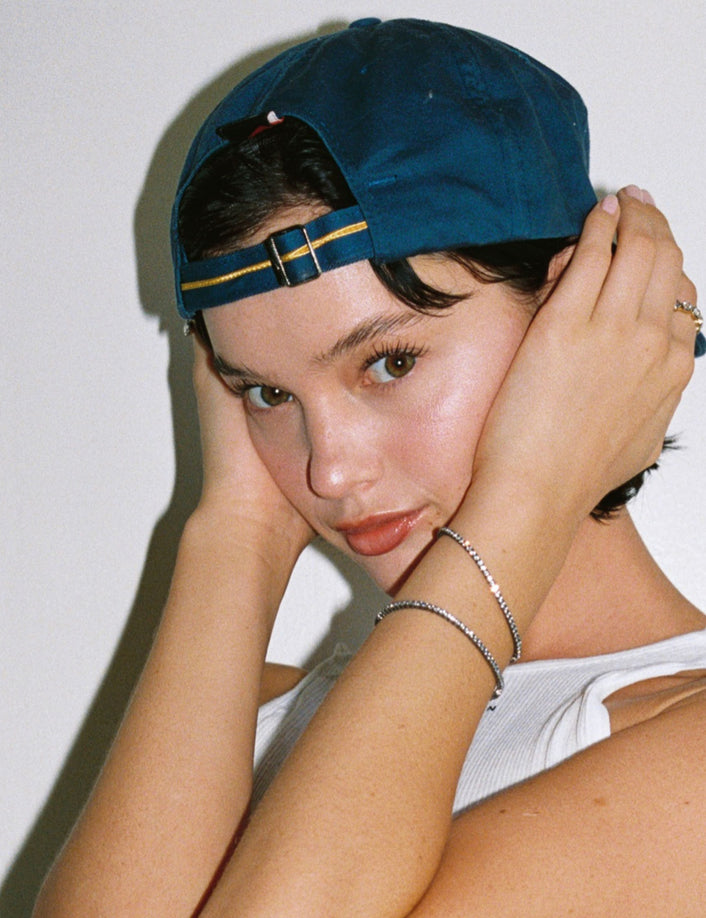 Model. Close up of woman in backwards blue hat with two gemstone tennis bracelets on white background. 