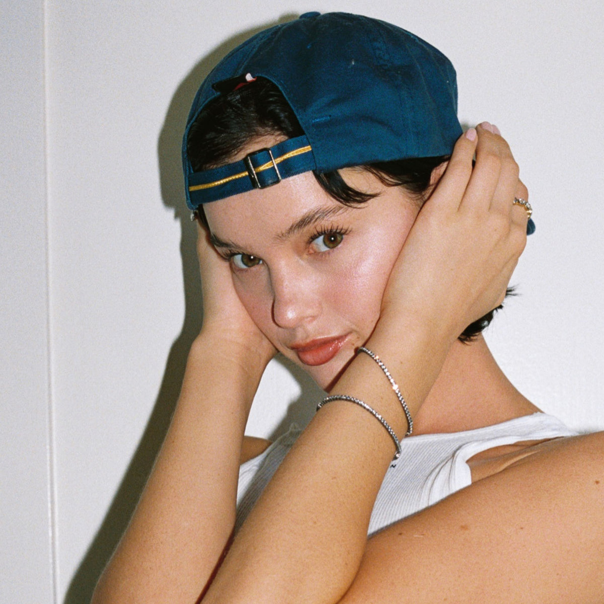 Model. Close up of woman in backwards blue hat with two gemstone tennis bracelets on white background. 