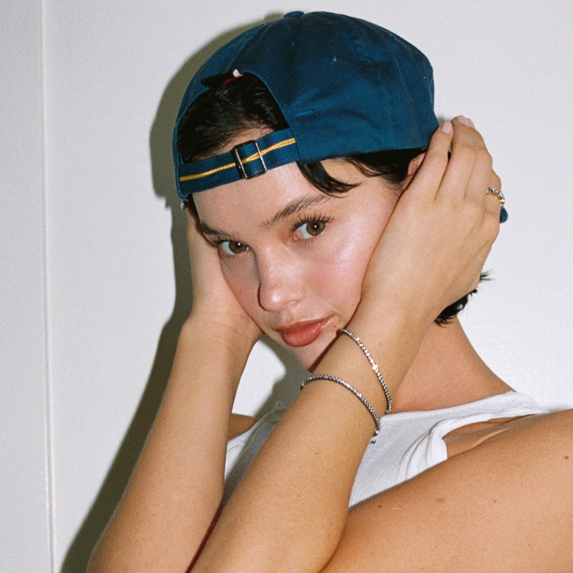 Model. Close up of woman in backwards blue hat with two gemstone tennis bracelets on white background. 