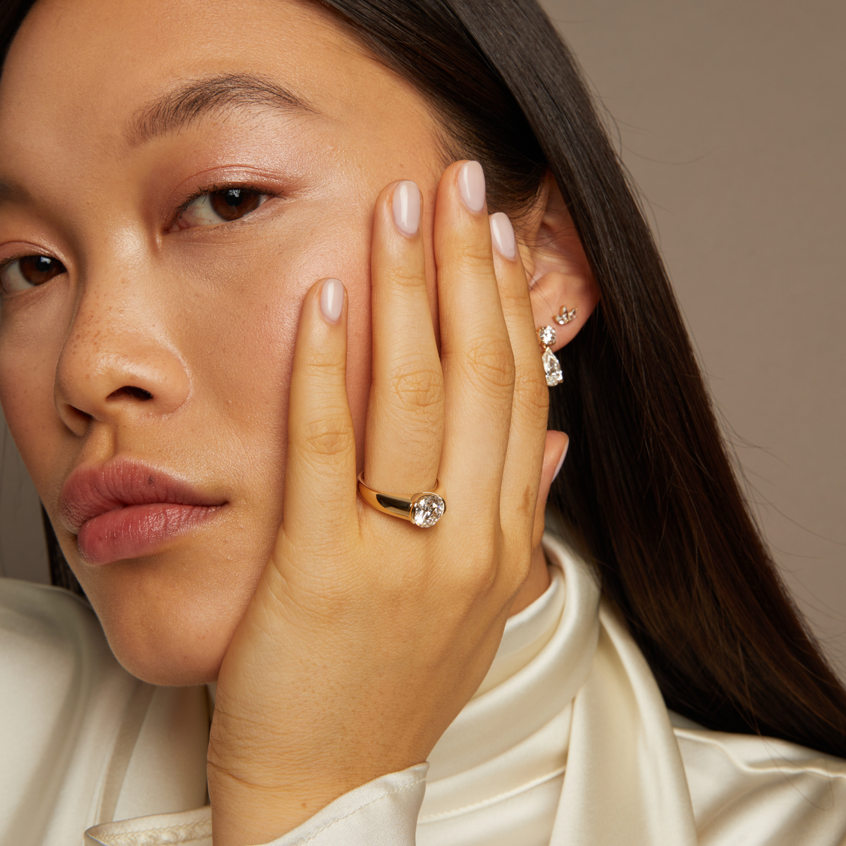 Model. Close-up of a woman wearing a gold ring and diamond earring against a neutral background