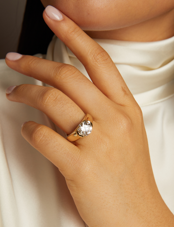 Close-up of a hand wearing a gold ring with a diamond on a neutral background