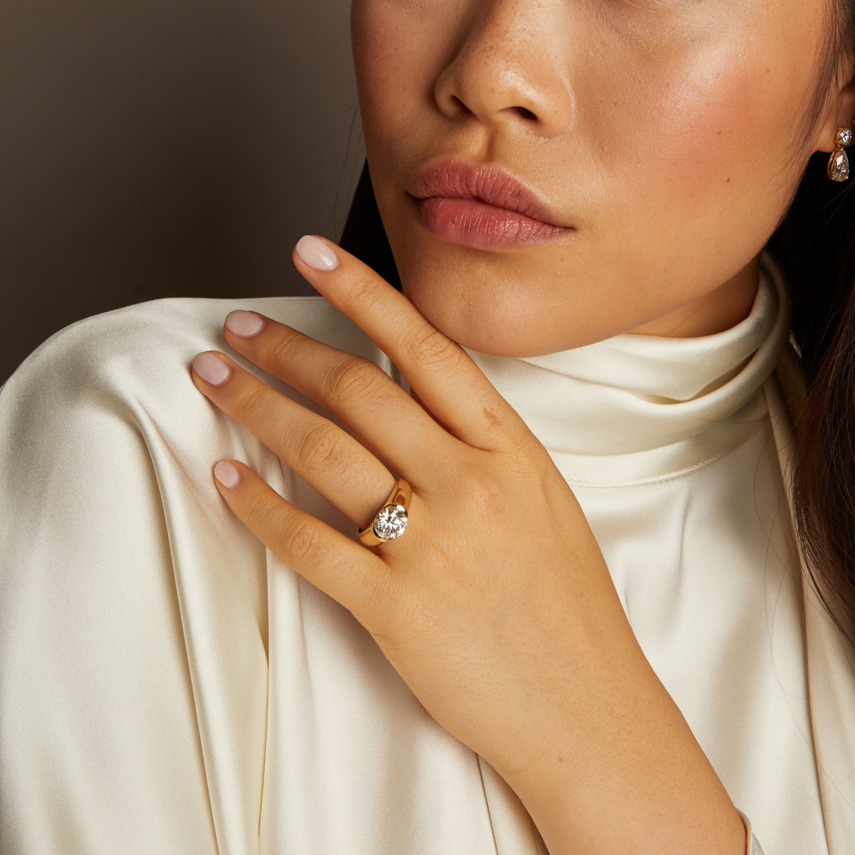 Model. Close-up of a woman's hand wearing a gold ring with a diamond, against a neutral background.