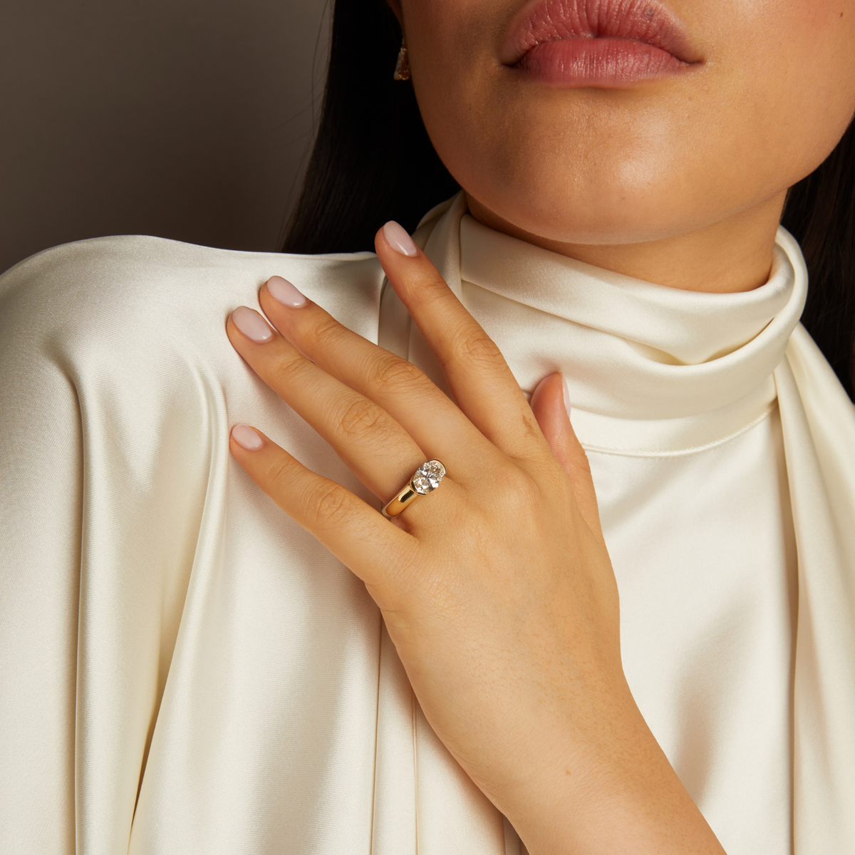 Model. Close-up of a hand wearing a gold ring with a diamond, against a neutral background.