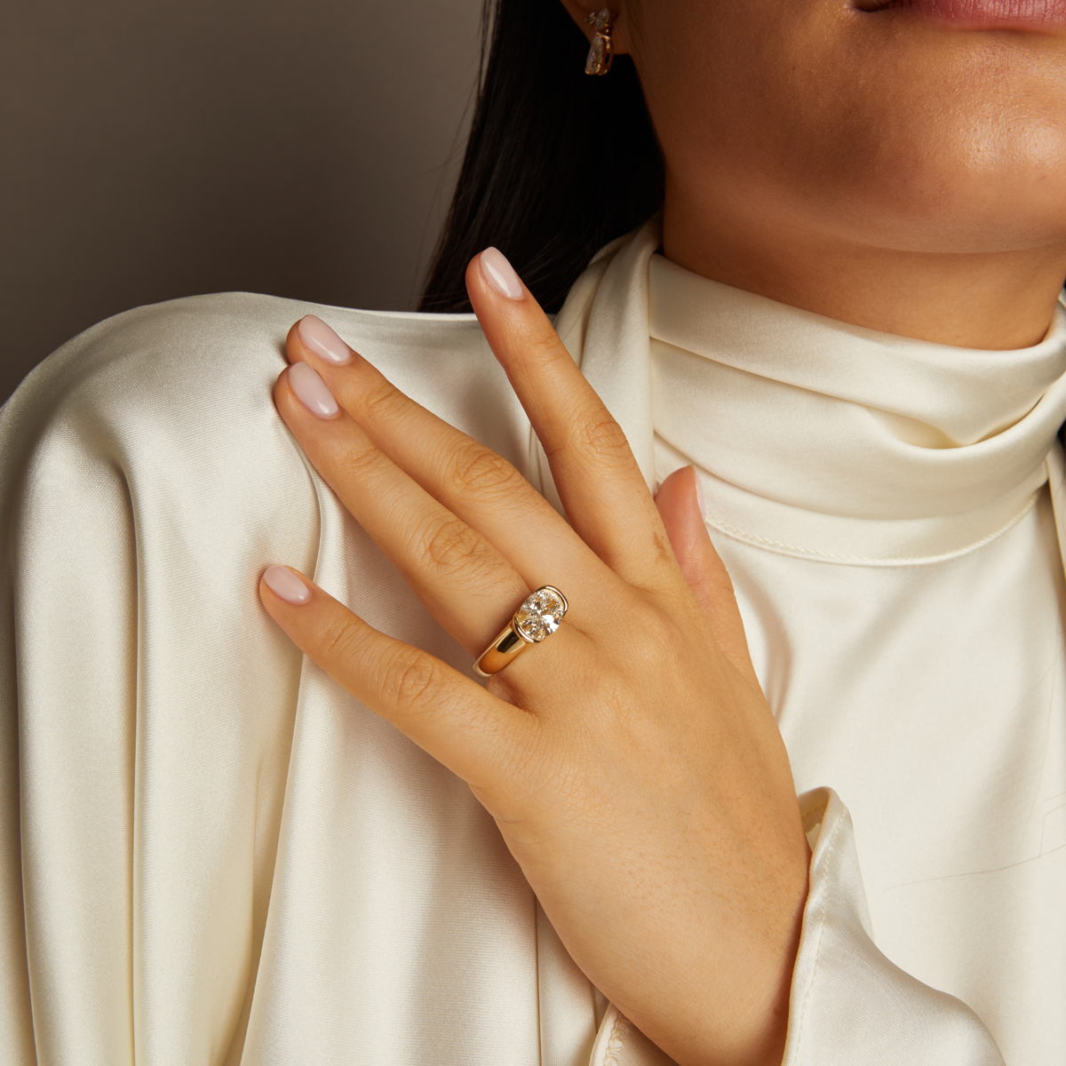 Model. Close-up of a hand wearing a gold ring with a diamond, against a neutral background.