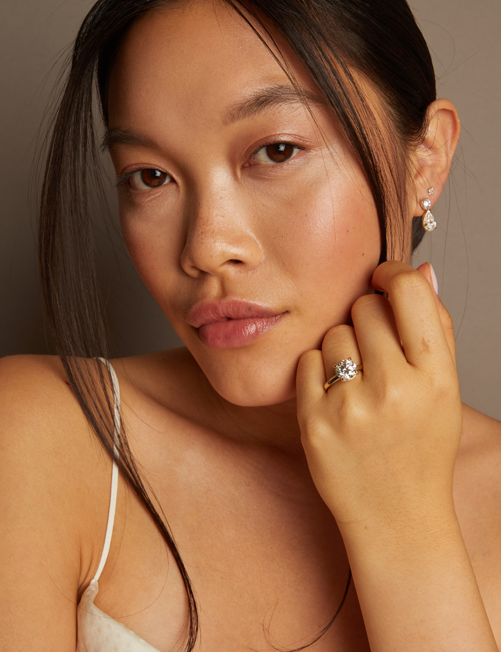 Model. Woman wearing earrings and a ring, posing with hand near face against a neutral background