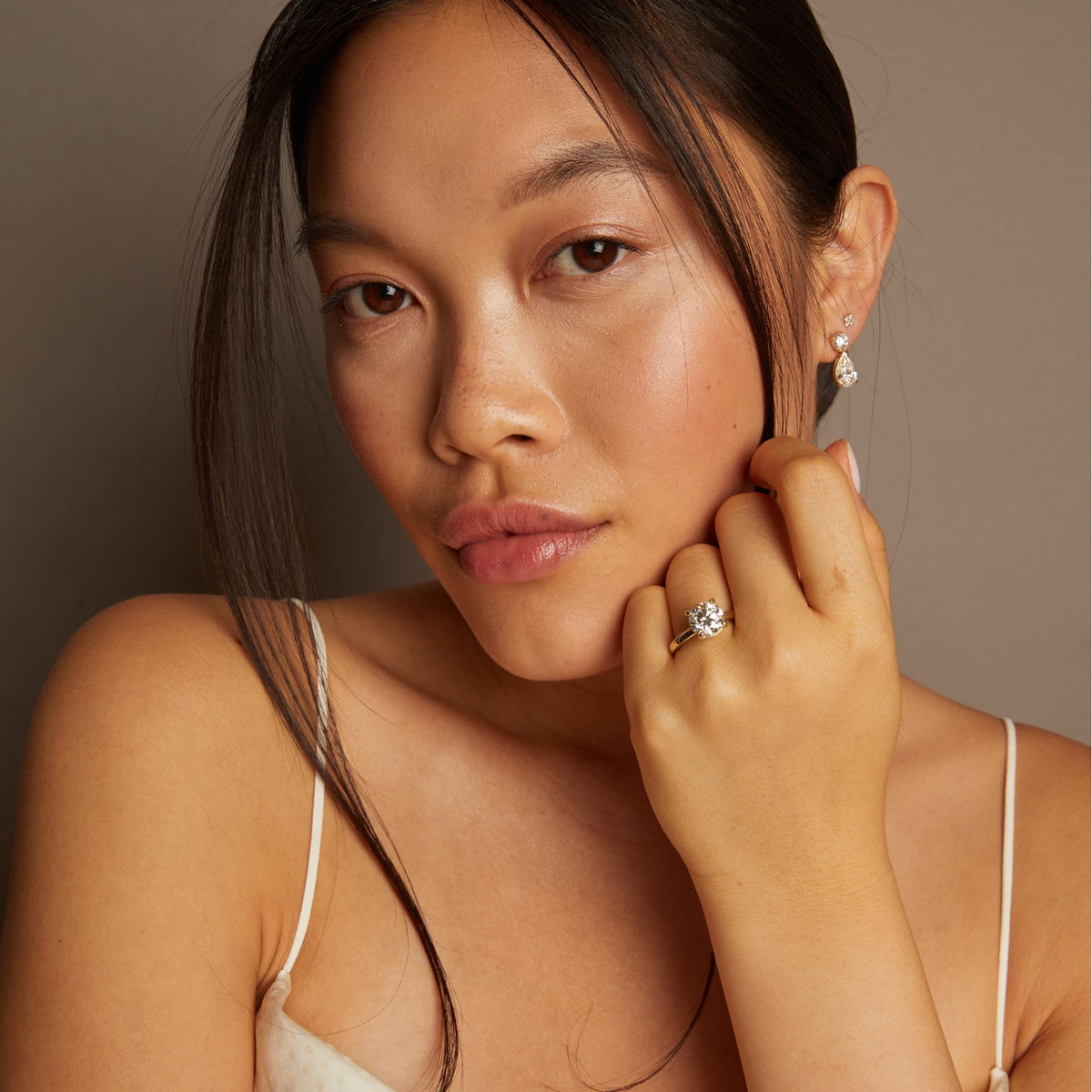 Model. Woman wearing earrings and a ring, posing with hand near face against a neutral background