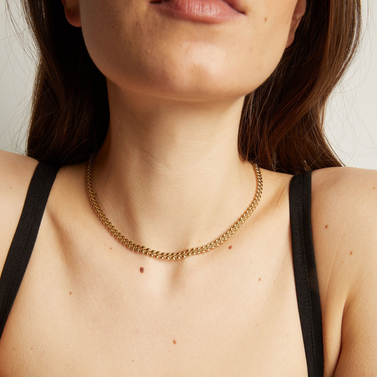 Model. Close up of a person wearing a gold chain necklace against a neutral background.