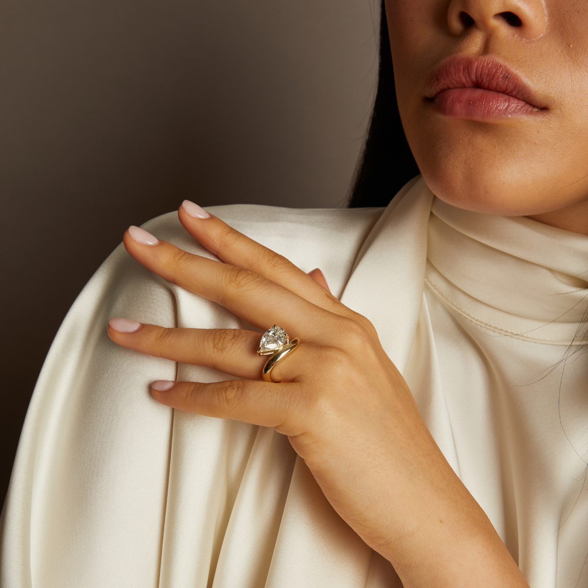 Model. Close-up of a hand wearing a gold ring with a diamond, against a neutral background.