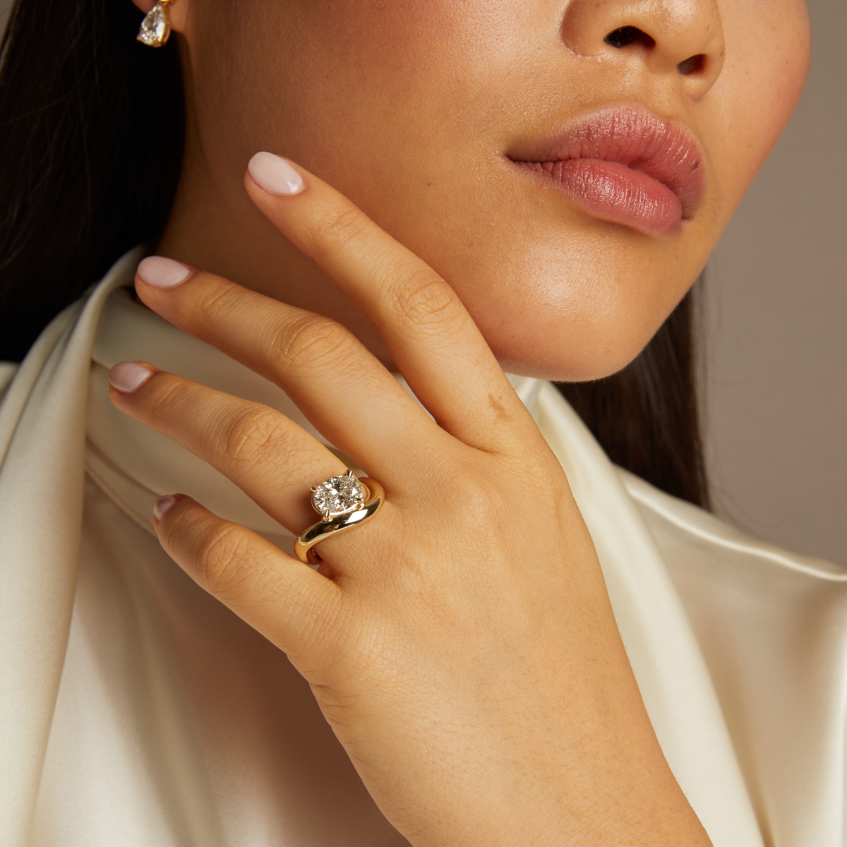 Model. Close-up of a woman's hand wearing a gold ring with a diamond, against a neutral background.
