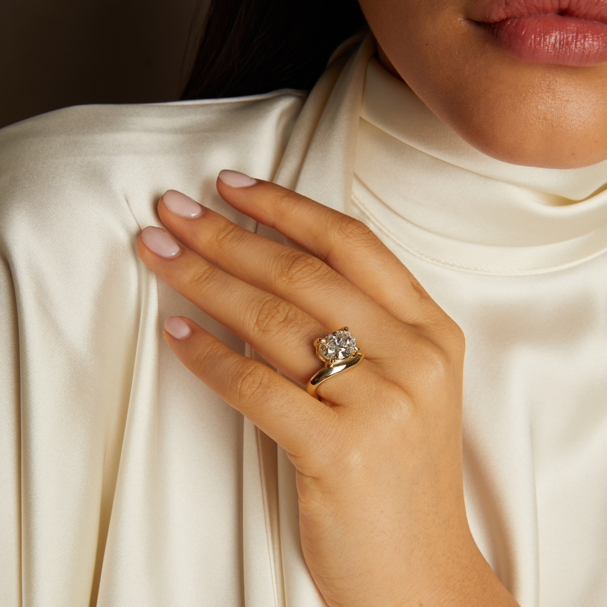 Model. Close-up of a hand wearing a gold ring with a diamond, against a neutral background.