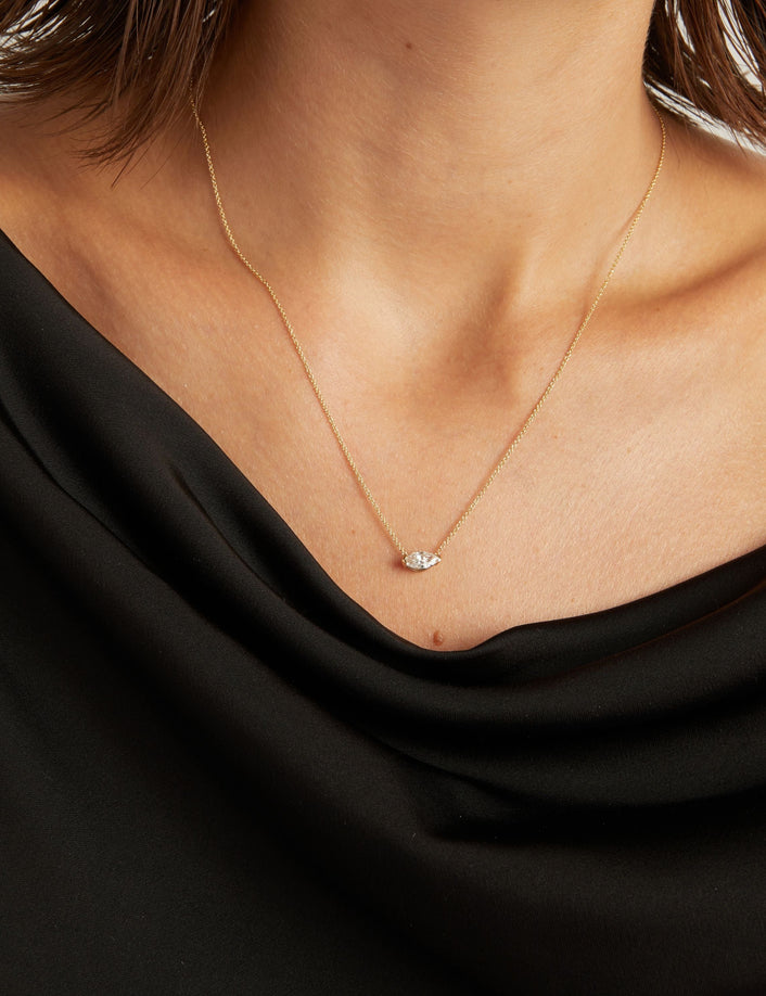 Model. Close up of woman wearing a black top with a marquise diamond solitaire necklaces on a beige background.