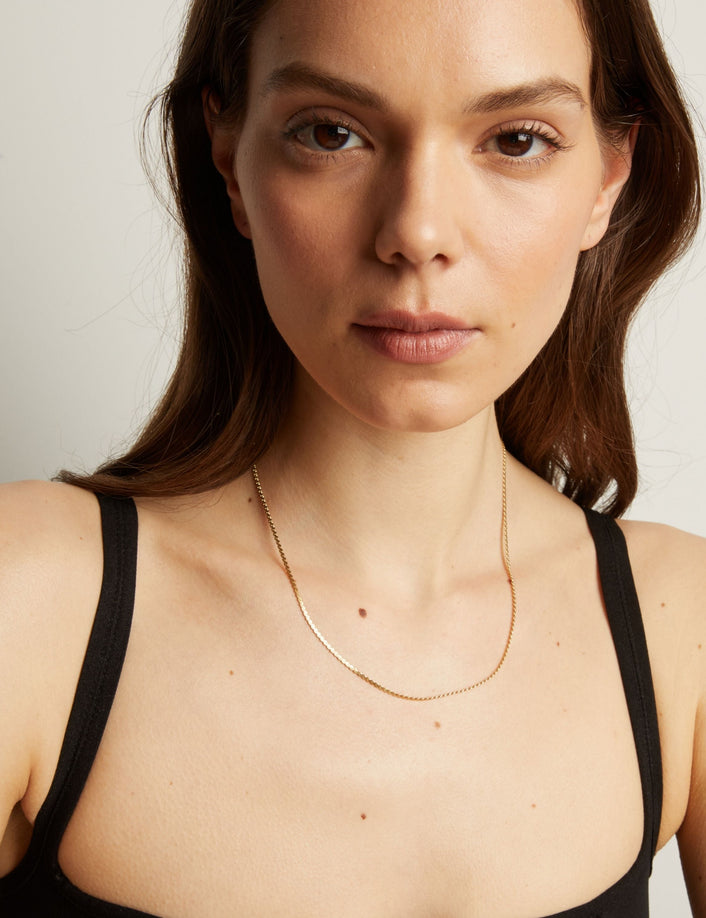 Model. Gold necklace on a woman wearing a black top against a neutral background.