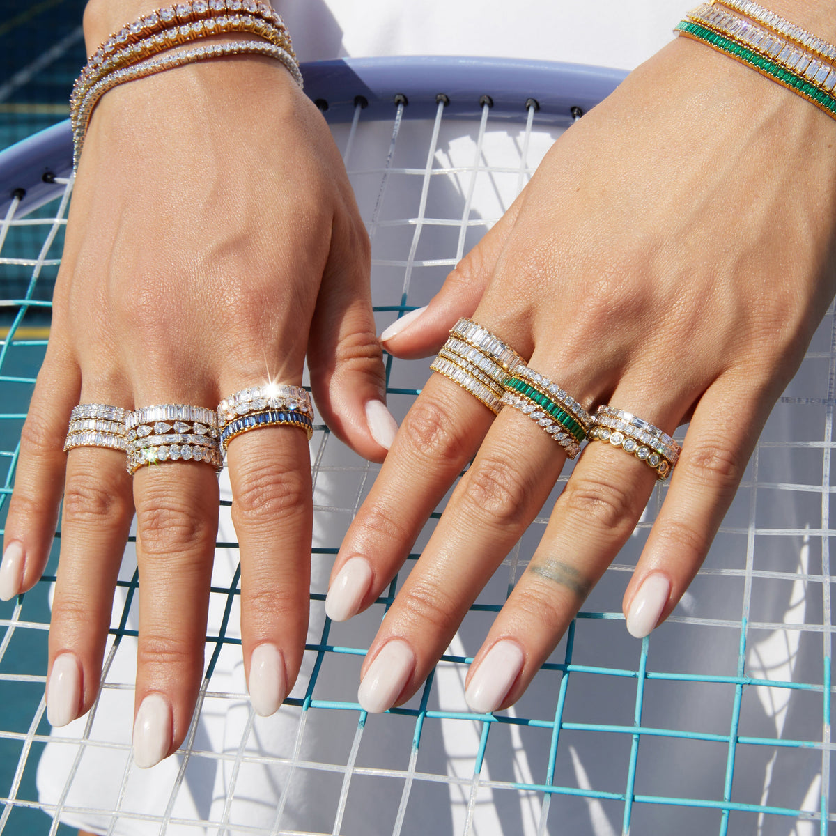 Lifestyle Model Close-up of a woman's hands resting on a tennis racket, wearing multiple gold rings and bracelets featuring diamonds, green, and blue stones