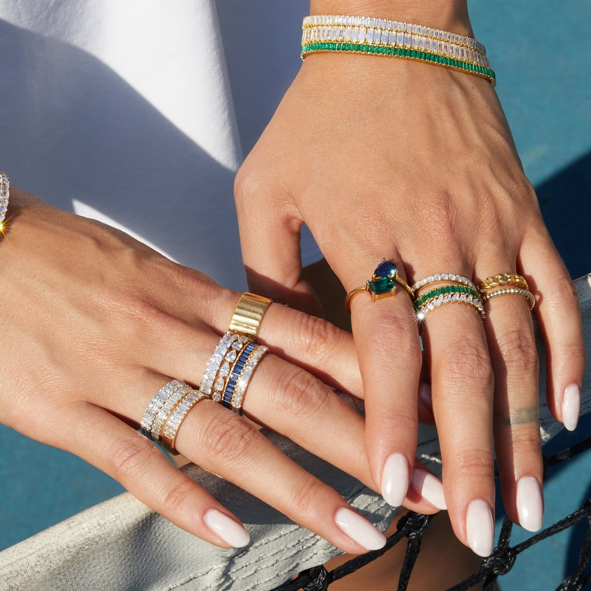 Lifestyle Model Close-up of a woman's crossed hands, wearing multiple gold rings and bracelets featuring diamonds, green, and blue stones
