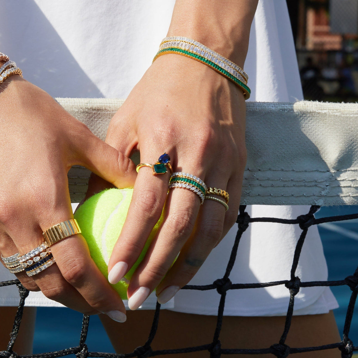 Lifestyle Model Close-up of a woman's hands holding a tennis ball against a net, wearing a gold two-stone ring with blue and green gemstones, stacked rings, and layered bracelets.
