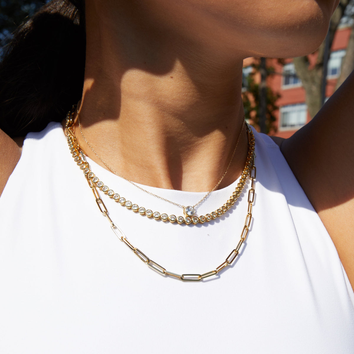 Lifestyle Model Close-up of a woman wearing a white top and three layered necklaces, including a delicate chain with a single round gemstone pendant, a diamond tennis necklace, and a gold paperclip chain.