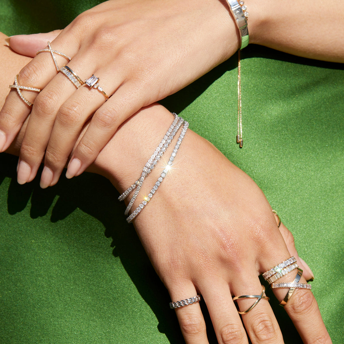 Lifestyle Model Close-up of a woman's hands resting against a green background, wearing multiple stacked gold and diamond rings alongside three layered diamond tennis bracelets.