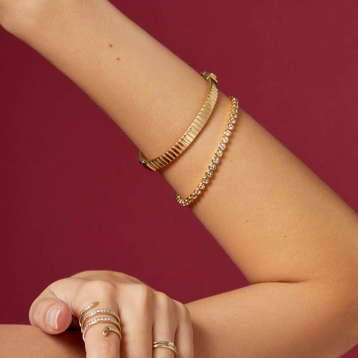 Lifestyle Model Close-up of a woman's bent arm against a dark red background, wearing a thick gold textured bangle paired with a single gold diamond bezel tennis bracelet on her wrist, and a curved, multi-band gold and diamond ring on her finger.